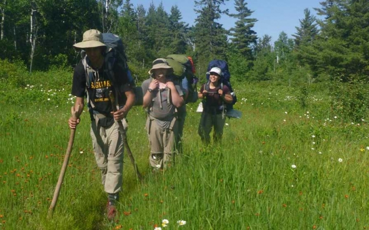 three students with backpacks hike in a grassy field